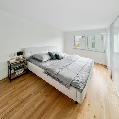 Gemütlich eingerichtetes Schlafzimmer mit großem Fenster, viel Tageslicht und Holzfußboden – Teil der Neubauwohnung in Satteldorf.