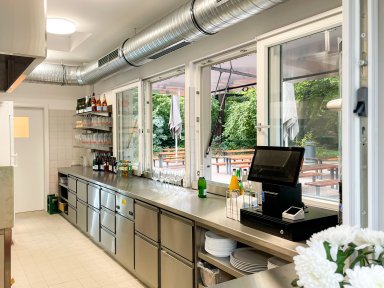 Innenansicht des neuen Kiosks KÖSK im Unteren Schlossgarten Stuttgart mit Edelstahltheke, Kühltheken, Kasse und geöffneten Ausgabefenstern zur Außengastronomie.