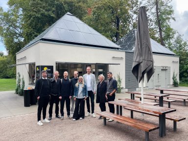 Acht Projektbeteiligte stehen gemeinsam vor dem fertiggestellten Kiosk KÖSK im Unteren Schlossgarten Stuttgart. Im Hintergrund sind die beiden markanten Pyramidendächer des Neubaus zu sehen.