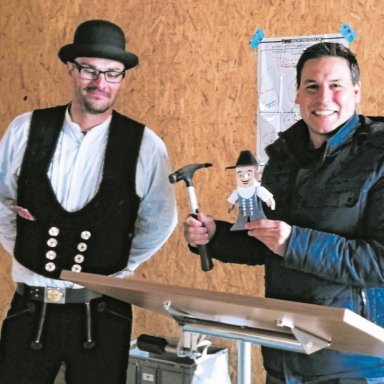 Zimmermann Achim Dausch von Bauer Holzbau überreicht Schnelldorfs Bürgermeister Tobias Strauß beim Richtfest der neuen Kindertagesstätte Schwalbennest einen Hammer und eine kleine Zimmermannsfigur. Im Hintergrund ist die Holzkonstruktion des Neubaus zu sehen.