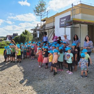 Zahlreiche Kindergartenkinder mit blauen Bauer-Holzbau-Kappen feiern gemeinsam mit ihren Erzieherinnen und weiteren Gästen das Richtfest des Kindergartenanbaus in Gröningen. Im Hintergrund ist der eingerüstete Holzbau mit einem Banner von Bauer Holzbau zu sehen.