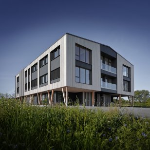 Modernes Verwaltungsgebäude von Bauer Holzbau in Satteldorf-Gröningen mit vertikaler Holzfassade, großen Fensterflächen und dunklen Fassadenelementen. Das Obergeschoss wird von schräg angeordneten Holzstützen getragen. Zwei verglaste Balkone und eine begrünte Außenanlage ergänzen die Architektur.